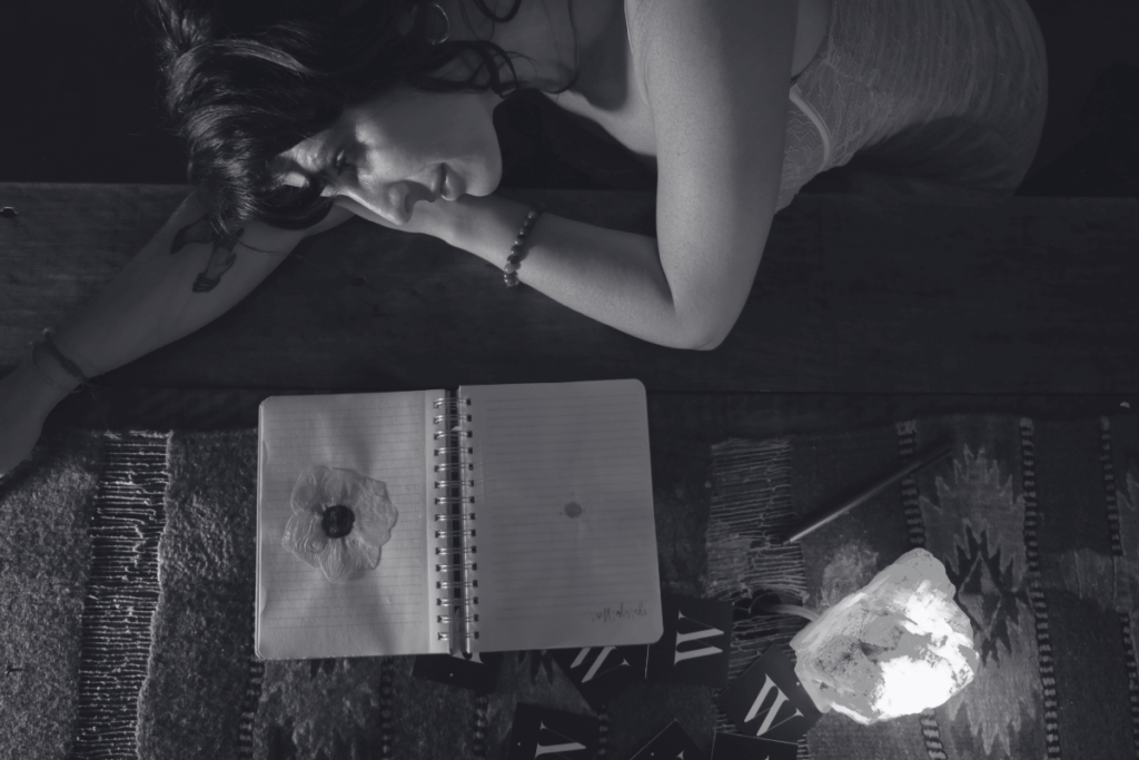 Wild Woman Haus® founder, Tristan Thibodeau, MS resting her head on a wooden table next to an open notebook and a glowing stone light