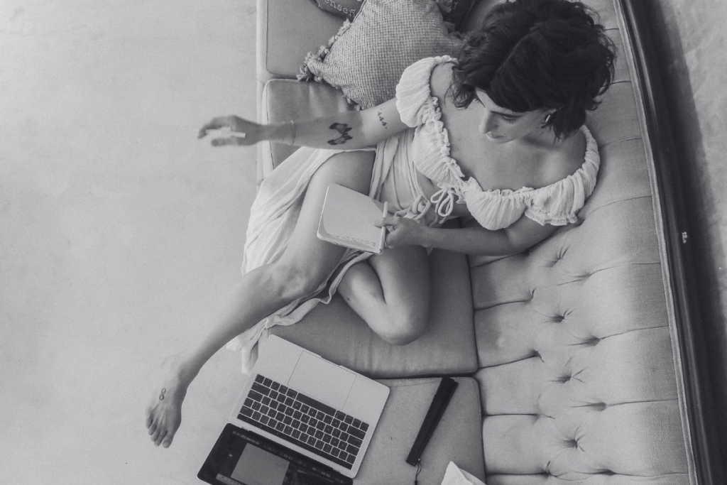Wild Woman Haus® founder, Tristan Thibodeau, MS sitting on tufted sofa in cream dress, with notebook and laptop